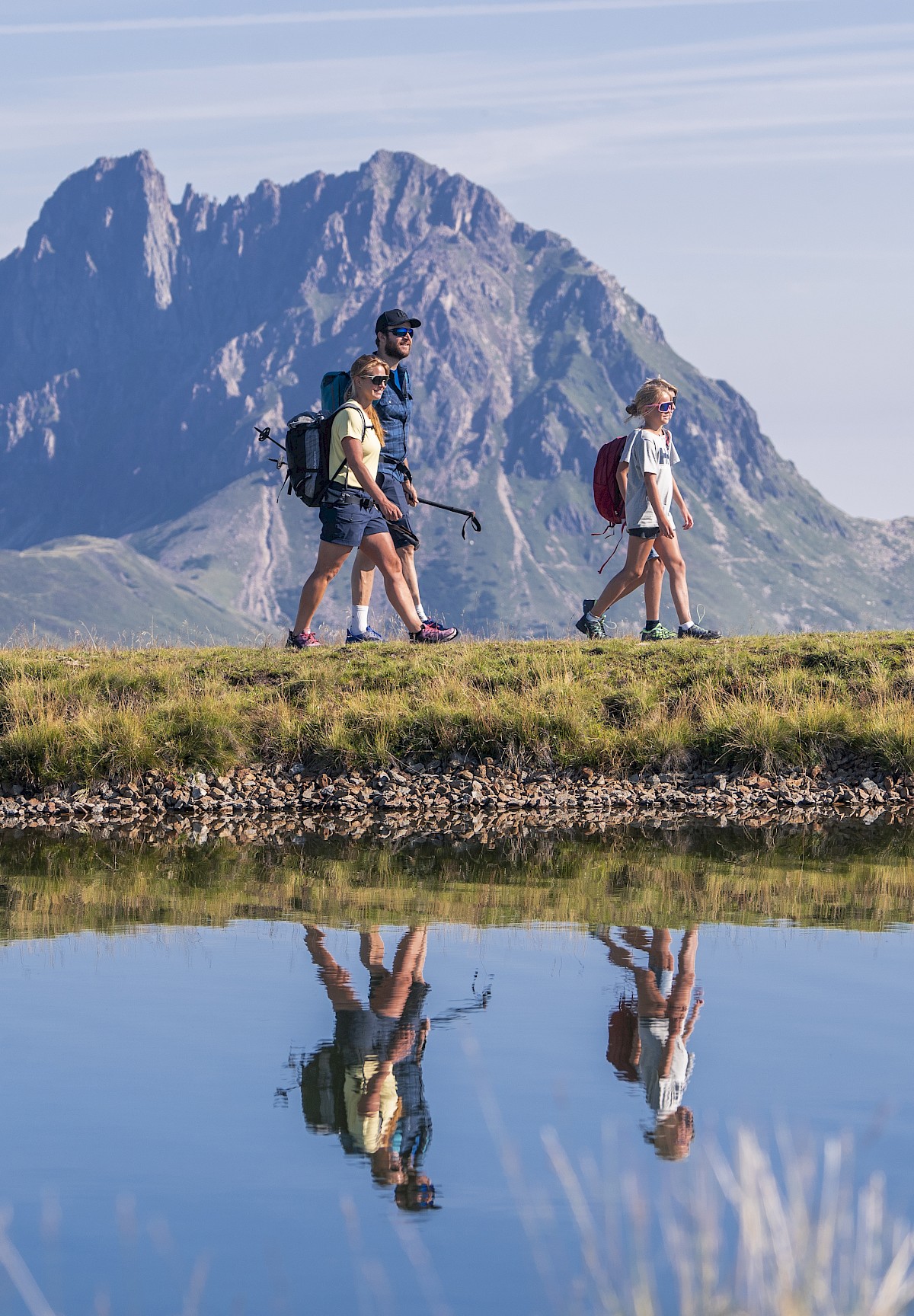 Eine Familie wandert bei Sonnenschein entlang eines idyllischen Speicherteichs im Höhenwandergebiet Wildkogel mit Blick auf den markanten Großen Rettenstein – perfekte Bedingungen für einen aktiven Sommerurlaub in den Bergen.