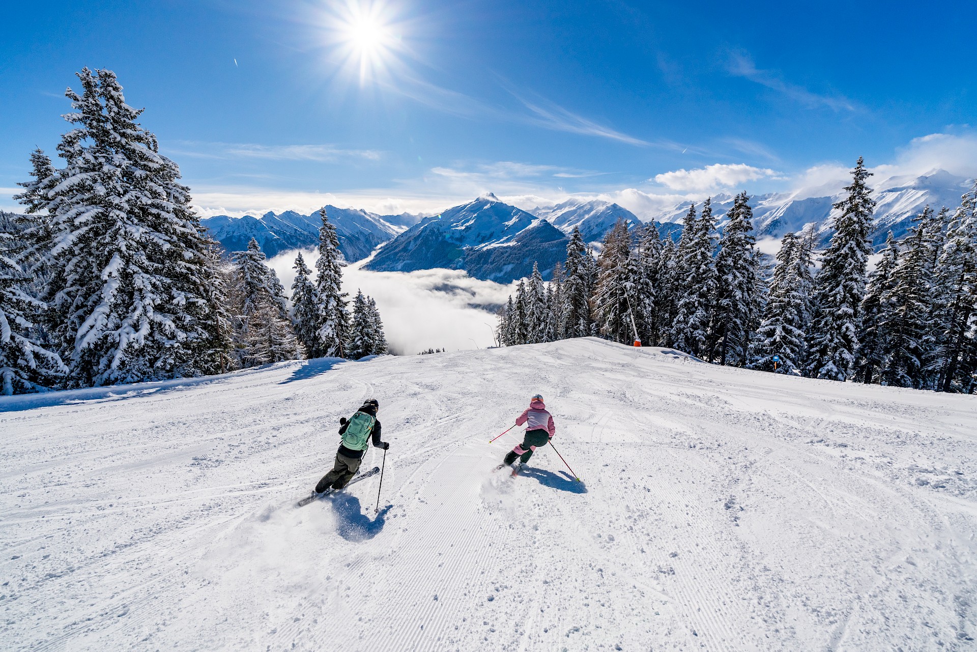 Zwei Skifahrer fahren eine frisch präparierte Piste in der Wildkogel-Arena im Salzburgerland hinunter, umgeben von schneebedeckten Bäumen. Die Sonne scheint strahlend vom blauen Himmel, während sich im Tal unterhalb Nebel sammelt. Im Hintergrund erheben sich beeindruckende, verschneite Alpenberge.