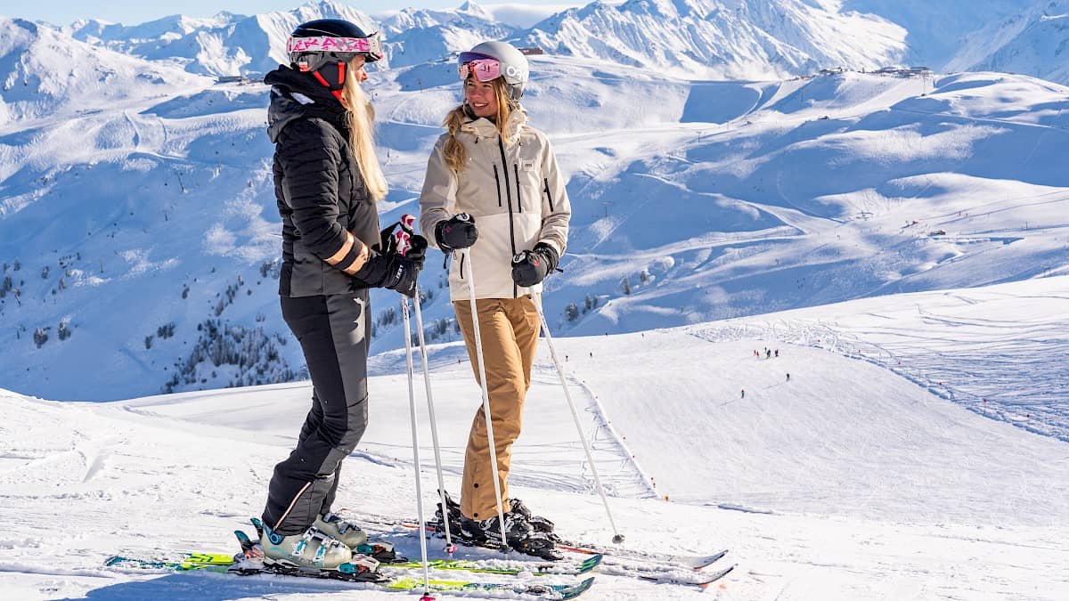 Zwei Freundinnen stehen in Skibekleidung bei blauen, wolkenlosem Himmel im Skigebiet und bewundern das Panorama.