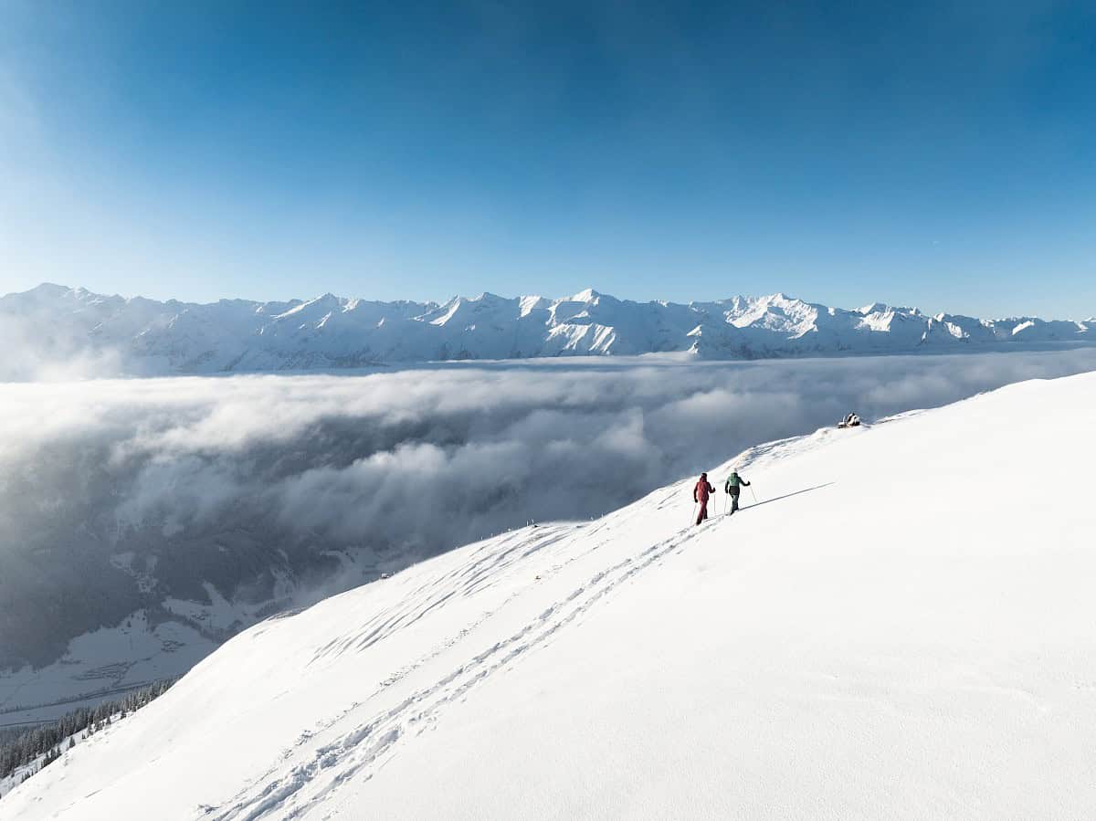 Zwei Schneeschuhwanderer ziehen ihre Spuren durch den frischen Pulverschnee am Wildkogel. Hoch über dem Nebelmeer genießen sie die Ruhe und den atemberaubenden Blick auf die verschneiten Gipfel der Hohen Tauern.