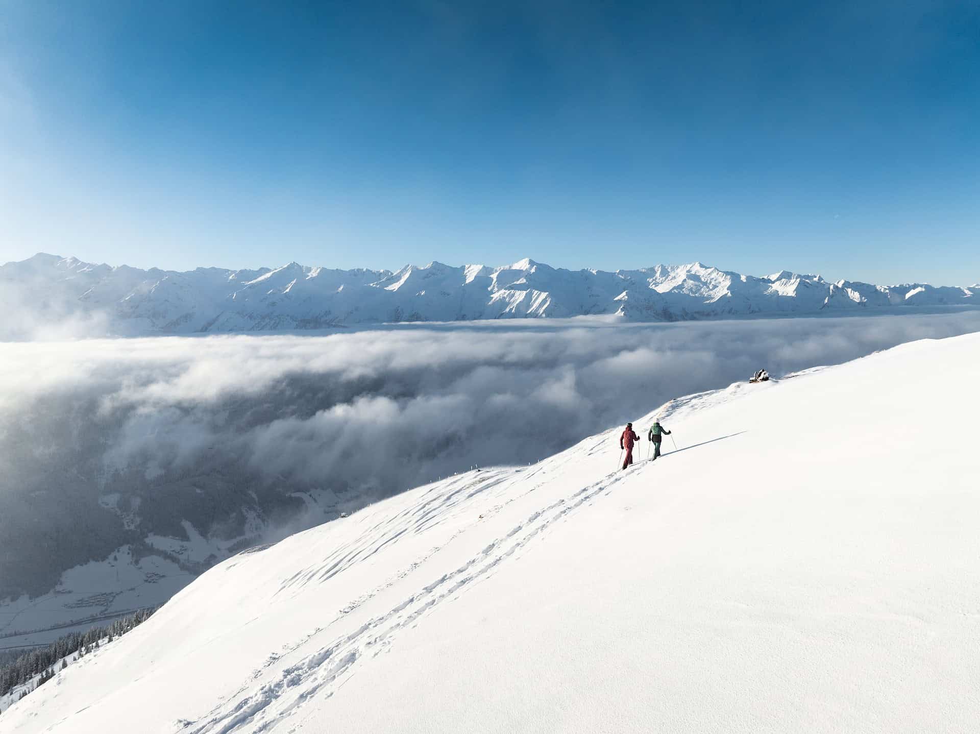 Twee sneeuwschoenwandelaars trekken hun sporen door de verse poedersneeuw van de Wildkogel. Hoog boven de mist genieten ze van de rust en het adembenemende uitzicht op de besneeuwde toppen van de Hohe Tauern.