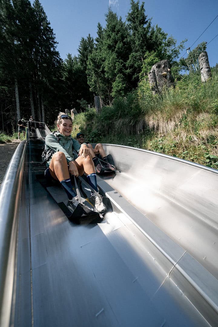 Zwei junge Erwachsene rutschen lachend auf einer Edelstahlrutsche im Wald entlang des Rutschenwegs in der Wildkogel-Arena – Sommerurlaub voller Abenteuer und Outdoor-Spaß in den österreichischen Alpen.