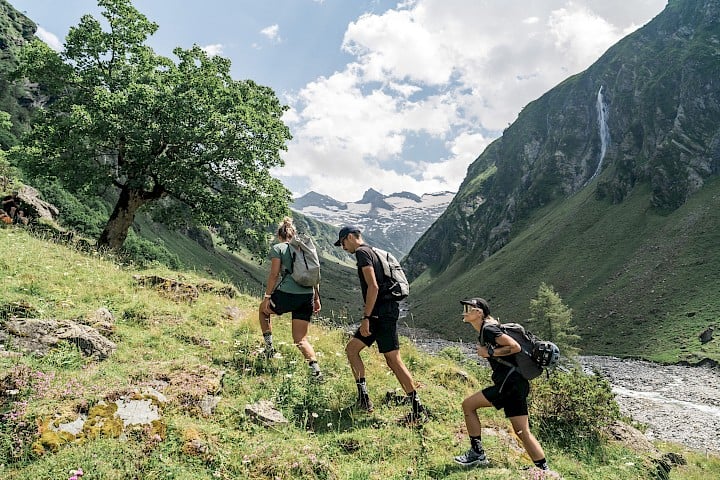 Drei Wanderer erklimmen einen Wiesenhang im Habachtal im Nationalpark Hohe Tauern – mit Blick auf Gletscher, Wasserfall und wilde Natur im Talschluss.