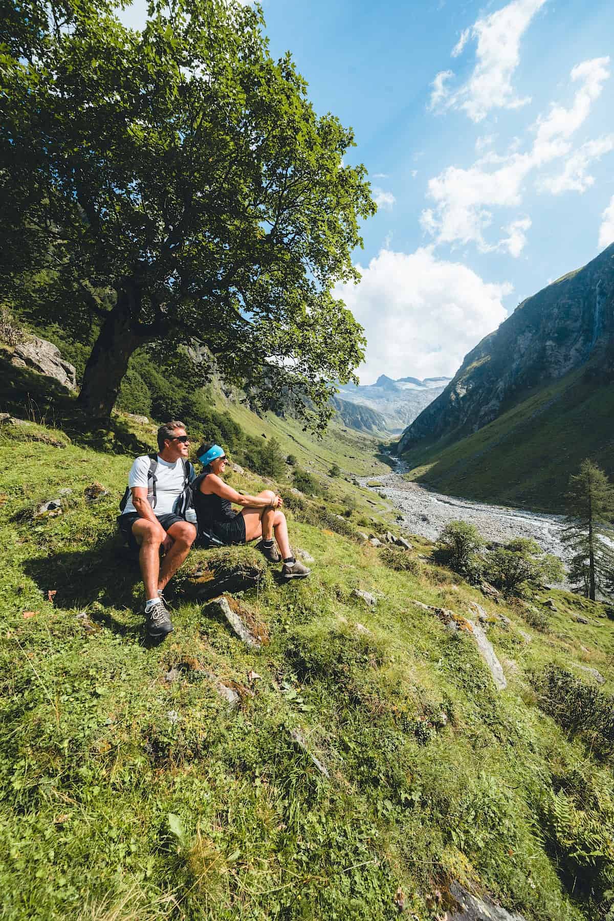 Ein Paar genießt eine Wanderpause unter einem schattigen Baum im Nationalpark Hohe Tauern mit Blick auf das wildromantische Habachtal – aktive Auszeit, Naturerlebnis und Erholung beim Wandern in der Ferienregion Wildkogel.