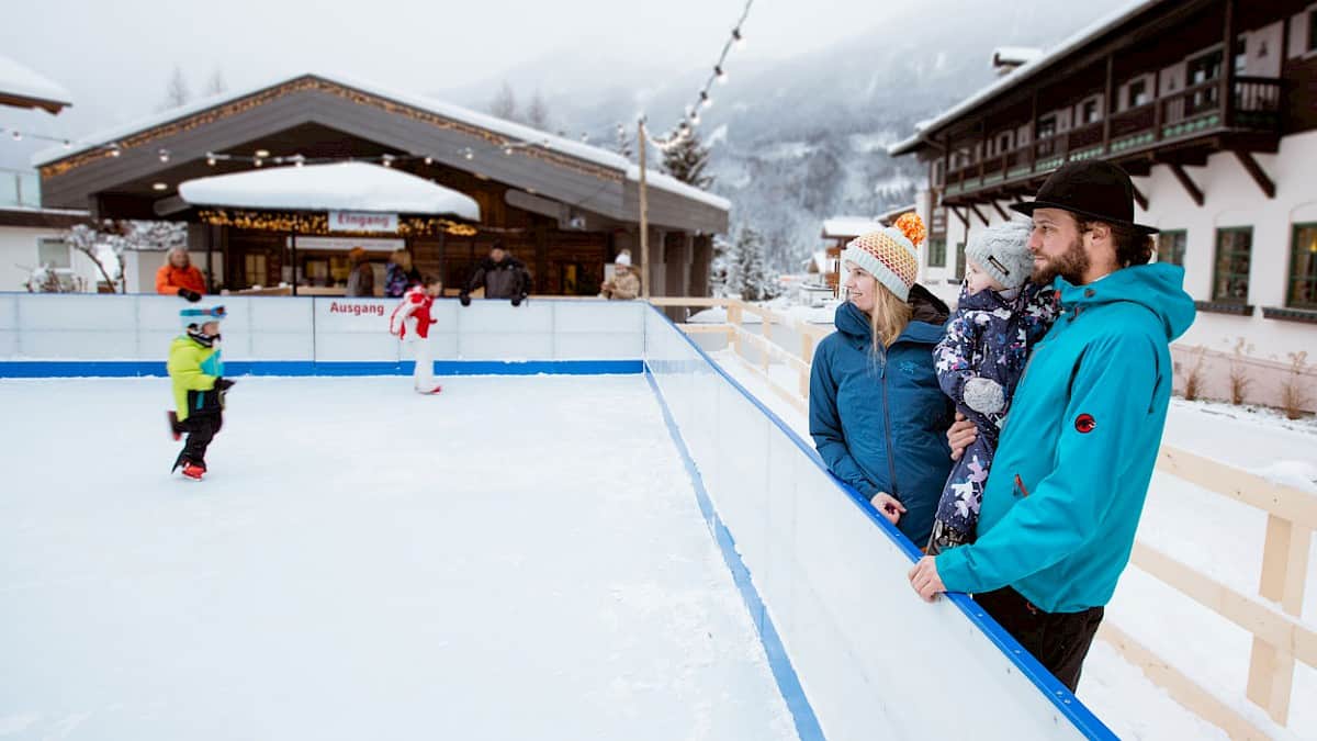 Wintervakantie met kinderen in Neukirchen – gezin kijkt naar het schaatsen op de ijsbaan in het dorpscentrum van de Wildkogel Arena.