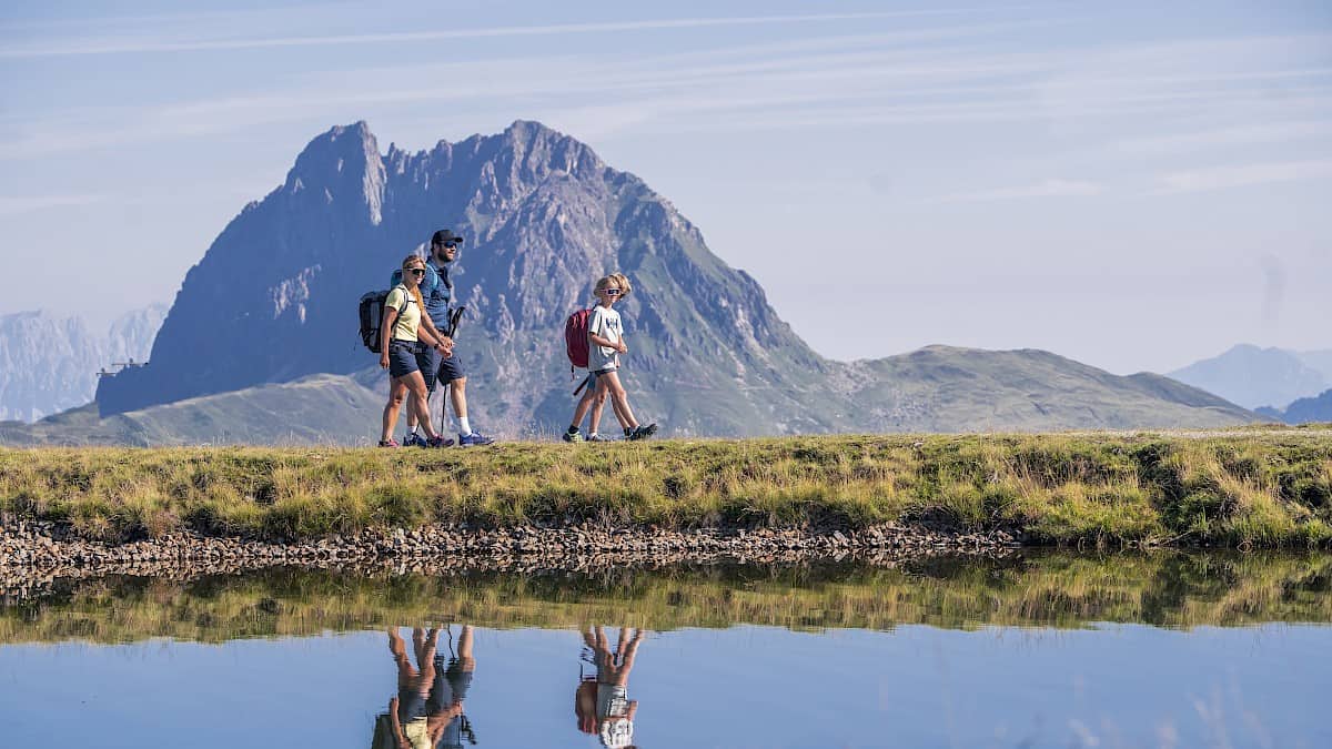 Rodina se za slunečného počasí prochází podél horského jezírka v oblasti vysokohorské turistiky Wildkogel s výhledem na impozantní vrchol Großer Rettenstein – ideální pro aktivní letní dovolenou v Alpách.