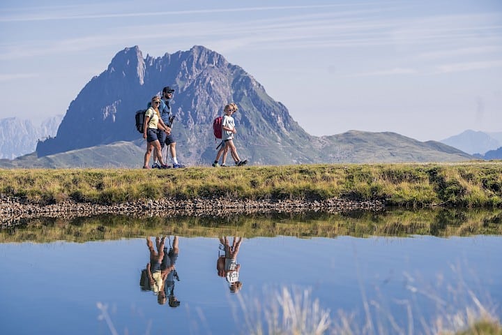 Eine wandernde Familie im Höhenwandergebiet Wildkogel, vor der imposanten Kulisse des Großen Rettensteins. Die klare Spiegelung im Bergsee verstärkt das Naturerlebnis beim Wandern in den Hohen Tauern – perfekter Sommerurlaub für die ganze Familie.