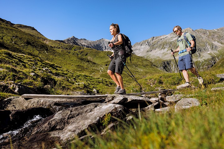 Zwei Wanderer überqueren auf einem Holzsteg einen Gebirgsbach in der hochalpinen Landschaft der Hohen Tauern – Sommerurlaub in den österreichischen Alpen voller Natur, Ruhe und Bewegung.